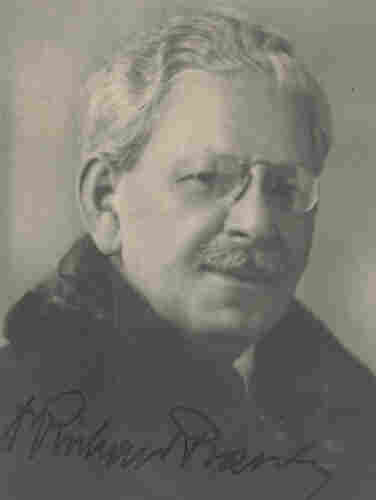 A portrait ID-style photo of a mature man with short grey hair. He is wearing frameless glasses. He is wearing a coat and a scarf.