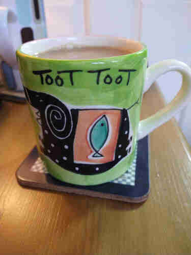 Mug of tea on a bedside cabinet, it has a green background and the words Toot Toot in black text at the top. 
In the middle is an odd looking black elephant with white spots, it is wearing an orange saddle with a green fish on it 