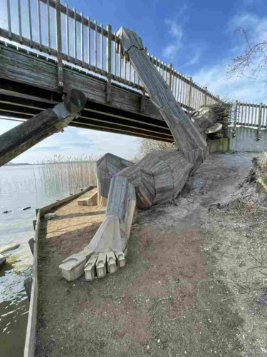 A large, wooden Thomas Dambo troll hiding underneath a bridge.