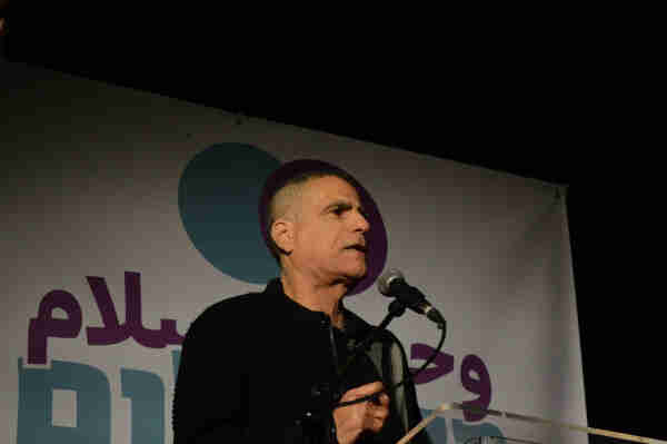 Mossi Raz speaking into a microphone, standing in front of a large sign that reads “Only Peace Will Bring Security” in Arabic and Hebrew.
