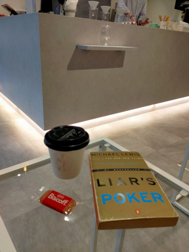 The paperback book is on a square glass table with metal edge. The book is gold with LIAR'S in big black letters & POKER in big blue letters  To the left is red plastic packaging of a Biscoff biscuit. In the center is a white paper coffee cup with a black plastic lid. In the distance is the barista's counter where white coffee filtered hand drip coffee drippers can be seen on top of clear glass carafes. The torso of the barista can be seen behind the counter, wearing a blue dress shirt
