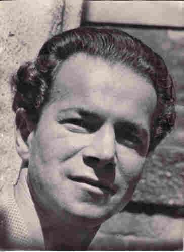 Photo of a man's face. His hair is pulled back with slight curls at the sides. There is a brightly lit face, but the left eye is in shadow. He is photographed against some kind of wall with visible lines. 