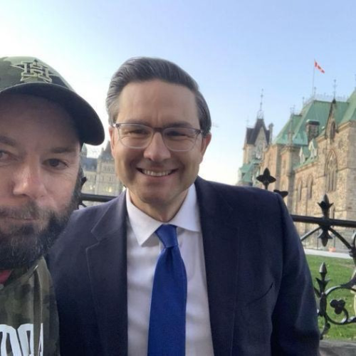 Poilievre grinning outside Parliament Hill. The individual taking the selfie has a beard and is wearing a baseball hat