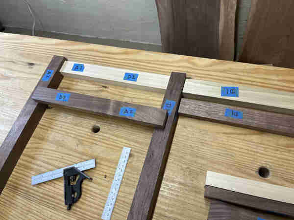 Dark brown and light colored pieces of wood laying on top of a workbench. They are aligned with the ends against each other in several different places at 90 degrees. There is blue tape marking the pieces.