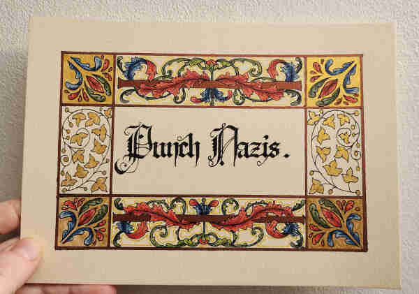 My hand holding a cream coloured piece of paper up against a white wall. It has the words 'Punch Nazis.' calligraphied in black ink, blackletter script with fancy majuscules, surrounded by a border of swirling acanthus leaves and flowery leafy shapes in gold, bronze, blue, red, and green.