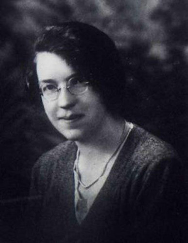 Black and white portrait of Jane Haining, posing with a slight smile, wearing glasses and a dark sweater with a necklace. Her hair is pulled back and she has a contemplative expression.