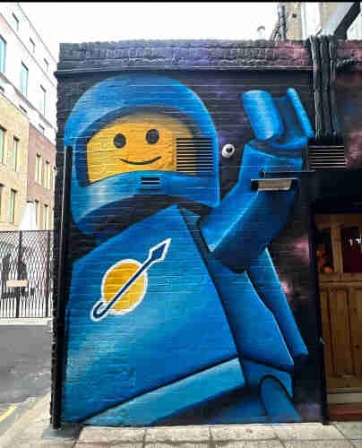 Streetartwall. A beautiful mural with a waving blue Lego man was sprayed/painted on a small brick wall. The background was painted black. The yellow head consists of two eyes and a smiling mouth. He has a yellow Lego Earth planet pictogram on his blue, angular body, wears a helmet and waves to the viewer with one arm. (There is a wooden door on the right-hand side and the road with a gate can be seen on the left-hand side of the mural).