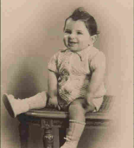 A photo of a baby boy sitting on an ornamented table. He has short hair and is smiling. He is wearing short sleeves and short trousers. 