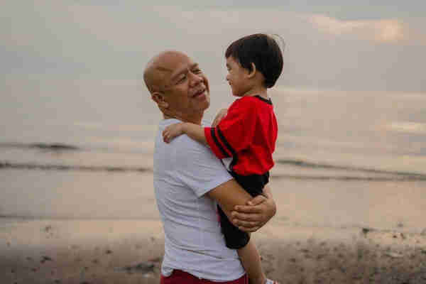 A father joyously holding his toddler-aged son to his chest against the backdrop of a hazy, blurred, and faded beach.