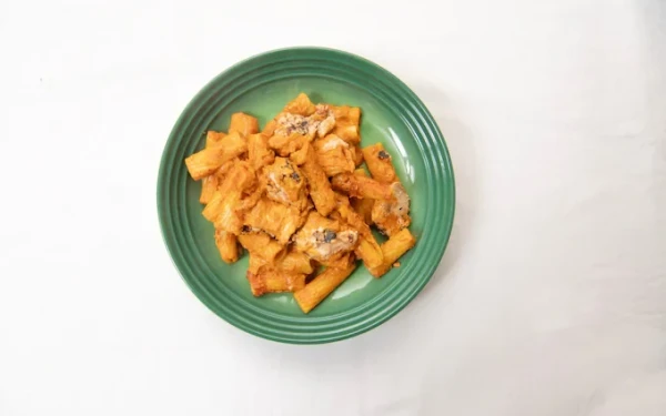 A green ceramic bowl on a white table containing tubular pasta shapes coated in an orange Chicken Tikka Masala sauce