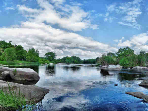 a small river, flowing peacefully through a green landscape, blue sky above with puffy, white clouds
