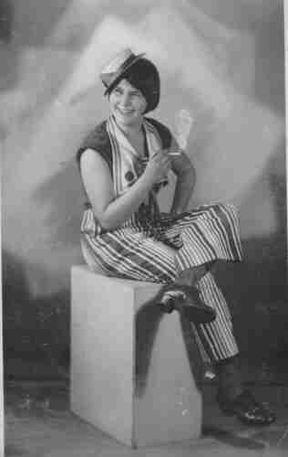 Studio portrait photograph of a young woman. She is seated on a cuboid with her leg overlapped. In one hand she holds a cigarette. She is wearing striped trousers and kennels with wide heels. Her shoulders are exposed. On his head is a tiny calico. She is smiling. 