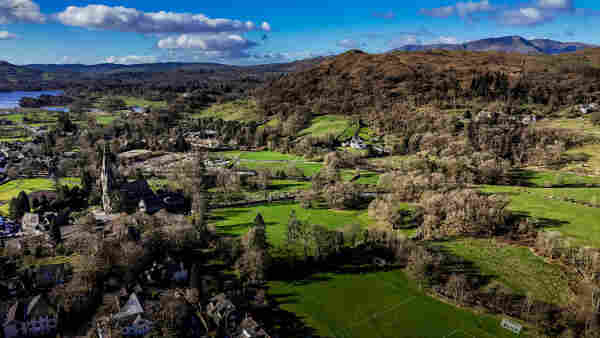 Aerial shot over the edge of the town of Ambleside in Cumbria, with tree covered hills and fields, lake to left frame and blue cloud scattered skies above