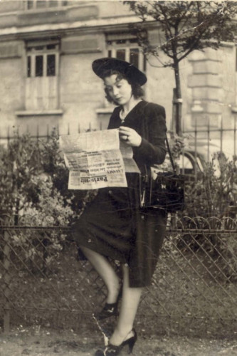 A woman in vintage clothing stands outdoors reading a newspaper. The attire includes a hat, jacket, knee-length skirt, and heels. The background shows trees and a building.

