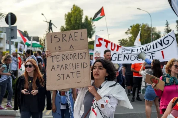 A woman holds a handwritten sign 'Israel is a terrorist apartheid state'. In the background is a banner saying 'Stop funding genocide!'.  [Etienne Torbey/AFP]