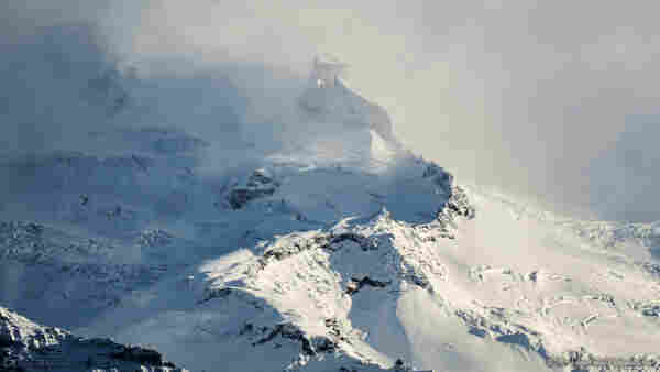 A photo of a mountain peak, covered in snow and ice. It's lit from the right, and deep crevasses are visible in the ice sheets near the summit. The background is obscured by mist and spindrift snow but at the centre of the shot is an unusual rocky stump. Behind it, and to the left, another slightly taller peak is visible.