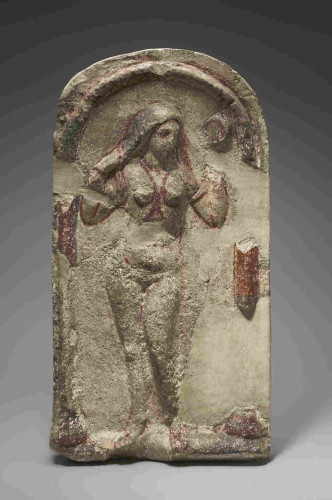 Limestone relief of Aphrodite in a niche, Roman artwork of 200-256 CE. The goddess is standing in the nude, wringing out her hair as if emerging from the bath or the sea. Red lines of paint suggest jewellery like a necklace and a body chain between the breasts.