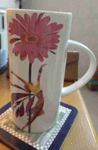 White mug on a bedside cabinet with handle to the right a large pink gerbera flower at the top and a leaping faerie below her arms are stretching up towards the flower, she is wearing a pink dress