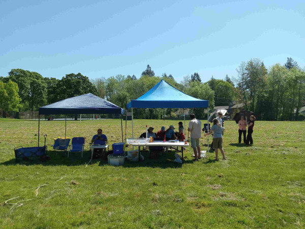 A grassy field with two pop-up canopies set up on it. Tables and chairs are under the canopies. Around a dozen people are milling about, getting model rockets ready to fly.