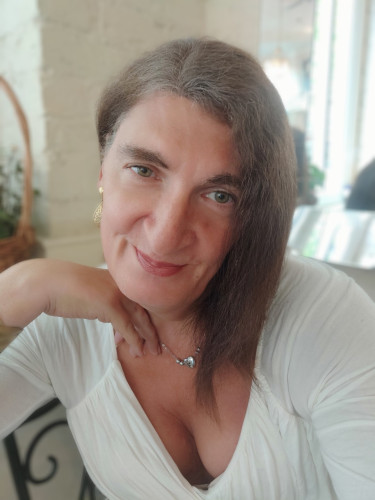 A selfie of a woman with frizzy shoulder length brunette hair resting her chin on the back of her hand. She's wearing a low cut white top showing some cleavage