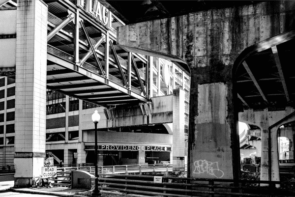A dynamic black and white photograph displaying the exterior of the Providence Place Mall with bold signage overhead. The image showcases a stark play of light and shadow, highlighting the geometric patterns of the bridge and building architecture. In the foreground, an empty street with a 'Road Closed' sign and a solitary street lamp adds a sense of desolation, while graffiti on a concrete pillar introduces an urban edge to the scene.