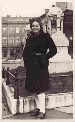 Black and white photograph of a woman. You can see her entire figure. Behind her a fenced-in statue and in the distance an apartment building. She is wearing a knee-length coat cinched with a belt, and a dotted headscarf on her head. She keeps her hands in her pockets. She is smiling.