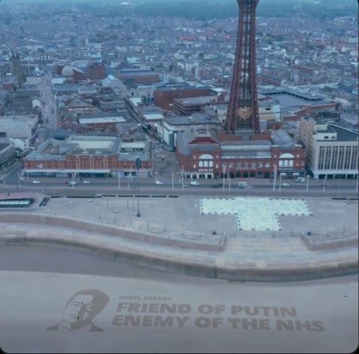 The seafront at Blackpool.  A portrait of Farridge has been made in the sand with the following text
NIGEL FARAGE
FRIEND OF PUTIN
ENEMY OF THE NHS