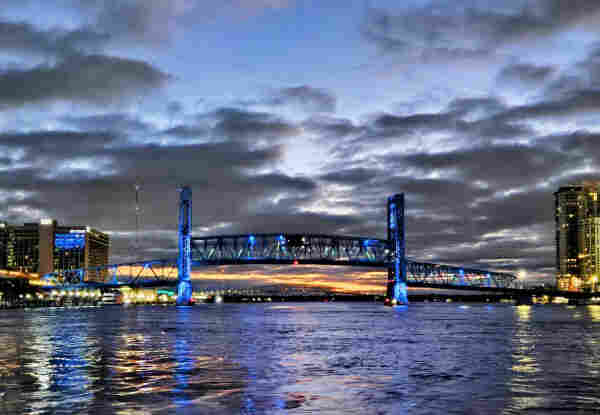Ominous clouds fill a dark blue sky above a wide blue river, spanned by a large metal bridge painted blue and illuminated with blue night lighting. Each shoreline is colorfully illuminated with tall buildings and their internal lighting, all reflecting onto the dark water below.
