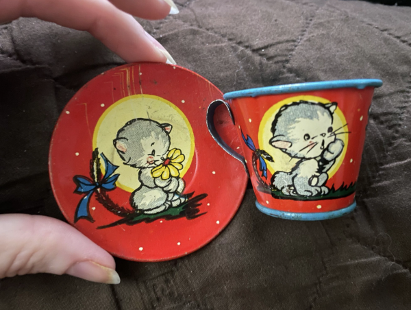 Very small red metal plate and cup covered with colorful graphics. The background is bright red and there are cute illustrations of a white faced grey kitten with a blue bow on its tail. In the saucer it is sniffing a yellow flower and it’s looking surprised with one paw up on the cup. My white, female fingers are holding the saucer for scale, showing it is less than two inches wide and the tea cup about an inch tall.