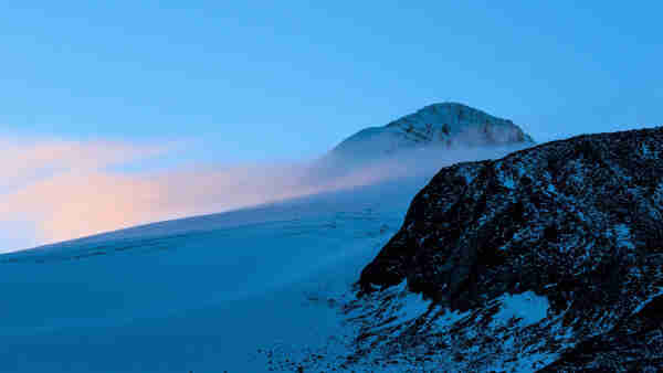 Blick hinauf über einen dunklen Felsen und mit Schnee bedeckten Gletscher zum Berggipfel. Links im Bild zarte pastellfarbene Nebelschwaden. 

View up over a dark rock and snow-covered glacier to the mountain peak. On the left in the picture, soft pastel-colored wafts of mist. 