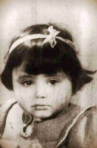 Picture of a little girl's face. She is dressed in a blouse buttoned up to the neck with ruffles on the shoulders. On her head is a headband with a bow. She has dark hair reaching her ears. 