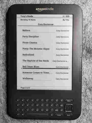 Photo of an Amazon Kindle device with integrated keyboard, showing a partial list of Cory Doctorow books.   