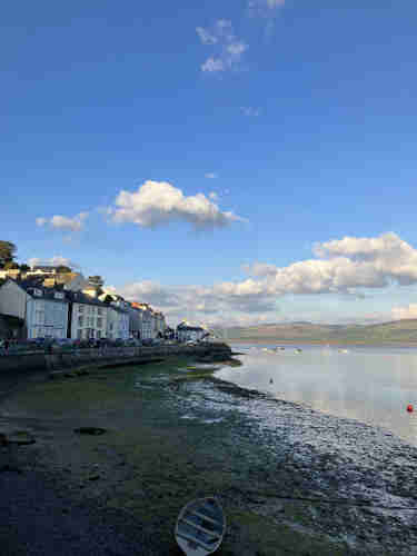 A coastal scene at Aberdovey with a row of houses by the water's edge, a blue sky with clouds overhead, and a small boat on the shore in the foreground.