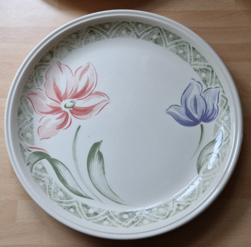A Staffordshire Tablewear dinner plate with a pink and a blue flower on either side. It has a green, sponge printed border of alternating triangles around the outer.