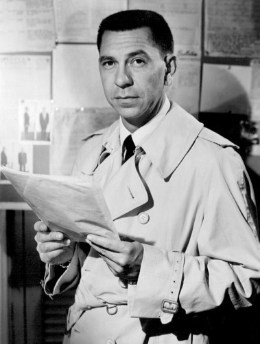 photo of Joe Friday holding a paper, wearing a trench coat. 