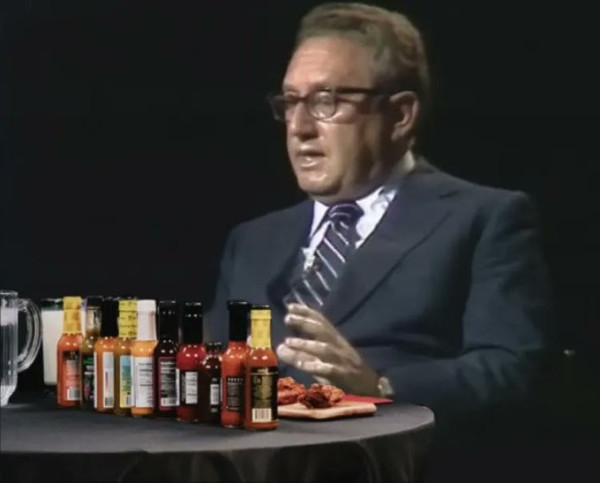 Still image. kissinger sitting in front of a round table with a black tablecloth, sweating like a mass murderer in the afterlife, a gauntlet of spicy wings and even spicier questions ahead of him. The wings have their respective bottled sauces behind them. 