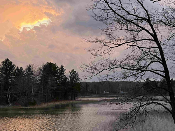 Photo includes our lake in the for and mid ground including the far shore lined with various trees. In the background is a cloud formation with sunlight beaming through on all shades of orange, fading into pink and the to purple. The spot where the dune shines through the brightness is on the left top of the photo. Hello to my blind friends! 