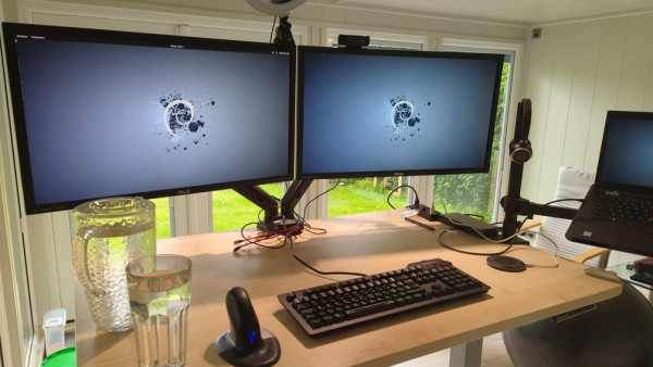 Photo of a plain wood desk, on which there are two monitors on an arm, connected to a hub, which is connected to a ThinkPad on its own flying arm / tray.

There is a keyboard and a mouse on the desktop, along with a jug of water and a glass.

The monitors all display the Debian logo, with a dark mode theme.

On top of the monitors there is a webcam, and a ring light. A pair of headphones is balanced on the pole for the laptop shelf.