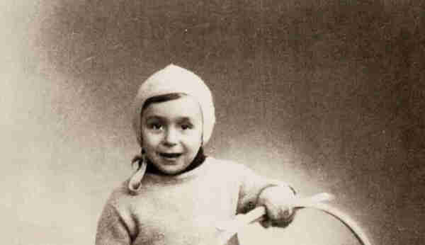 Photo of a young boy wearing a cap covering his head. He is wearing a light-coloured jumper. In his left hand he is holding a toy - a circle.