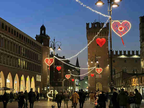 A vibrant evening scene in a bustling city square adorned with festive lights. Heart-shaped illuminations create a romantic atmosphere, hanging from lamp posts and strung across the open space, glowing warmly against the twilight sky. Historic buildings line the square, their traditional architecture and lit windows adding to the ambience. People, bundled in winter attire, stroll through the area, some pausing to admire the decorations, while the soft glow of street lamps casts a cozy light over the entire scene.