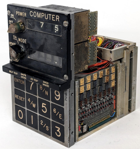 A compact aerospace computer with a numeric keypad and digit displays on the front. A circuit board with multiple flat-pack ICs is visible.