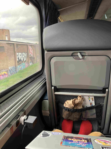 View from a train seat with ample leg room. A laptop is on my knee and the brown paper bag in the seat back pocket has 2 cans of Bell’s two hearted ale in it. Outside a nondescript brick wall with graffiti on it.
