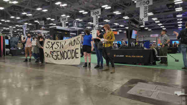 Protesters stand in a row in front of the US Army booth inside of an Expo center. The banner reads SXSW Runs On Genocide. 