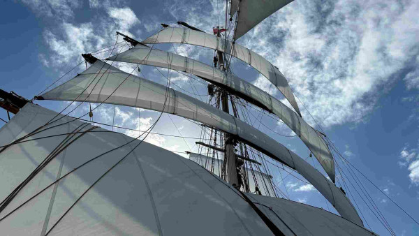 Four square sails set and one still furled on the yard. Two people are barely visible through the foot of the top gallant and upper top sail. 