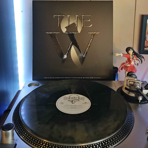 A Gold & Black Galaxy vinyl record sits on a turntable. Behind the turntable, a vinyl album outer sleeve is displayed. The front cover shows the Wu-Tang "W" logo as a shadow. Text on the cover says "THE W". 