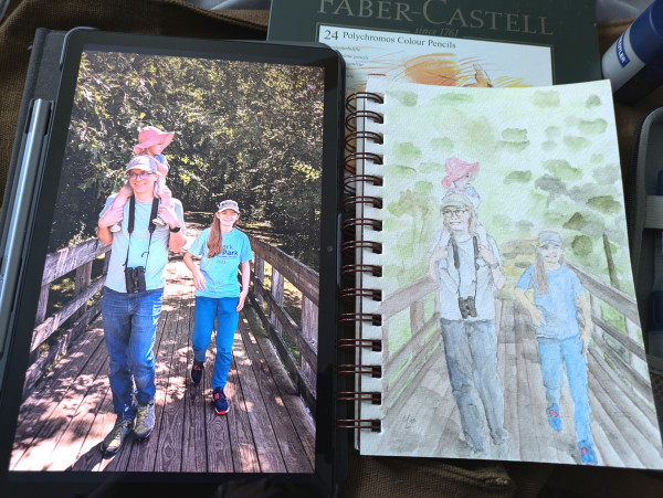 A watercolor painting of a male walking with a teenage child and a toddler on his shoulders. They are on a wooden bridge on a path in the woods. Next to the painting is the reference photo displayed on a tablet and some art supplies are visible in the background.
