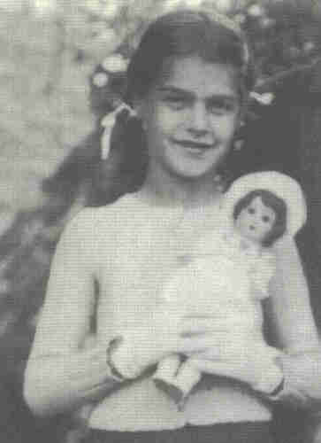 Young girl in jumper. In her folded hands she holds a large doll. There are sleeves on her hands. Her dark hair is pinned up in a crouch. She is smiling.