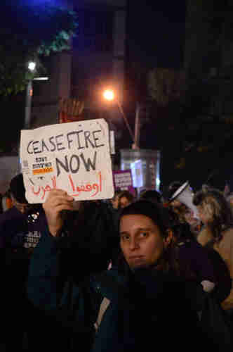 Night time, street lights, crowd of people. A woman holding a sign above her head that reads: “Ceasefire Now” in English and Arabic.