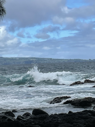 A blue green white capped wave coming onto a black sand beach with rocks. The sky has grayish blue clouds that are broken to reveal some blue sky.