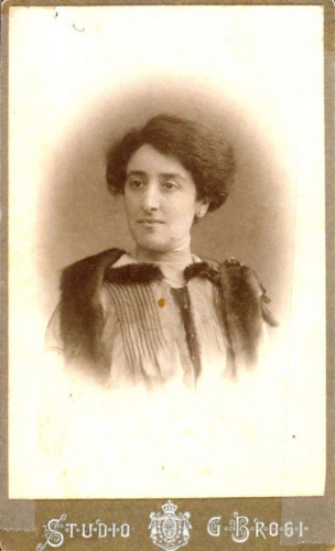 This image depicts a vintage portrait of a woman  in formal attire, featuring a high-necked blouse with ruffled details and a dark, wide ribbon tied at the neck. The person is styled with an upswept hairstyle typical of the late 19th or early 20th century. The portrait, likely a cabinet card based on its style and format, is marked with the text "Studio G. Brogi" at the bottom, indicating the photographer or studio's name. The background of the photo is plain, focusing attention on the subject and their attire.